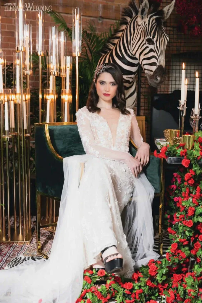 In this image from Mariage Elegant's vendor listing for Canada, Studio Caroline Théoret, under the wedding beauty category, showcases a bride elegantly seated in a luxurious setting. The bride is wearing a stunning, long-sleeved lace wedding gown with intricate beadwork, featuring a deep V-neckline and a sheer tulle overlay. Her dark hair is styled in soft waves adorned with a delicate headpiece, complementing her polished makeup. She is seated on a plush green velvet chair surrounded by vibrant red roses and golden candelabras, adding a touch of opulence. A zebra stands behind her, creating a unique and exotic backdrop that adds a whimsical element to the composition.