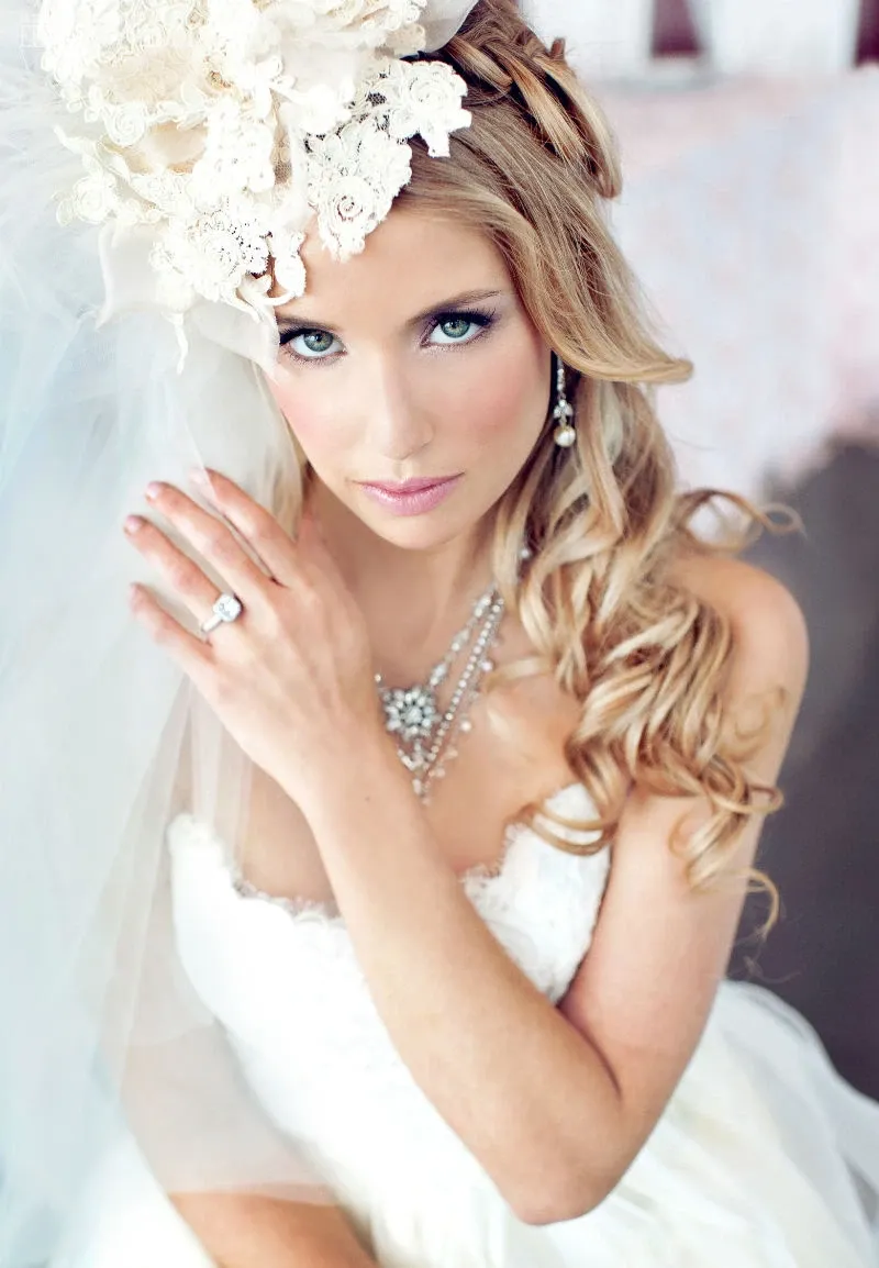 The image showcases a stunning bride with long, curled blonde hair styled elegantly by Studio Caroline Théoret, under the Wedding Beauty category. She wears a beautifully intricate white lace headpiece adorned with floral designs, complementing her flowing veil. Her makeup is flawlessly applied, highlighting her blue eyes and giving her a radiant glow. The bride is dressed in a classic white wedding gown, the fabric cascading gracefully. She delicately holds the veil with her left hand, showcasing a sparkling engagement ring. Her ensemble is completed with a sophisticated necklace and drop earrings, emphasizing the elegance typical of a Mariage Elegant event.