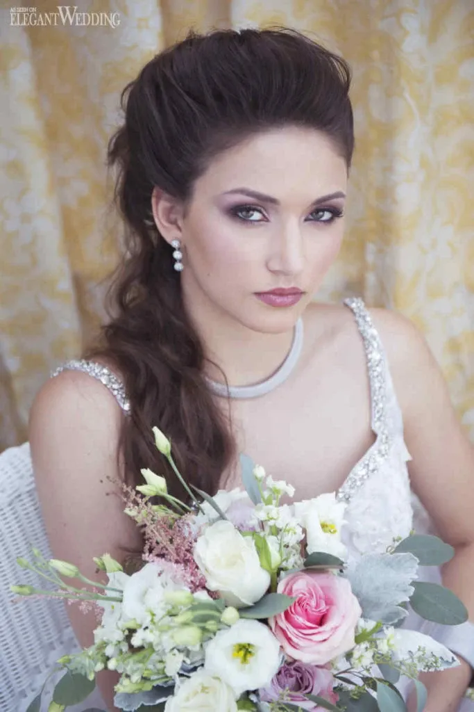 In this elegant portrait by Studio Caroline Théoret, a wedding beauty vendor with Mariage Elegant in Canada, a bride is seated against a soft floral backdrop. She wears a sparkling, bejeweled gown that complements her sophisticated jewelry, including pearl drop earrings and a delicate necklace. Her makeup features a classic, subtle smokey eye and a soft, pink lip color, enhancing her graceful appearance. Her hair is styled in an intricate yet natural updo. In her hands, she holds a beautiful bouquet composed of pale pink roses, white blooms, and lush greenery, capturing the essence of wedding beauty and sophistication.