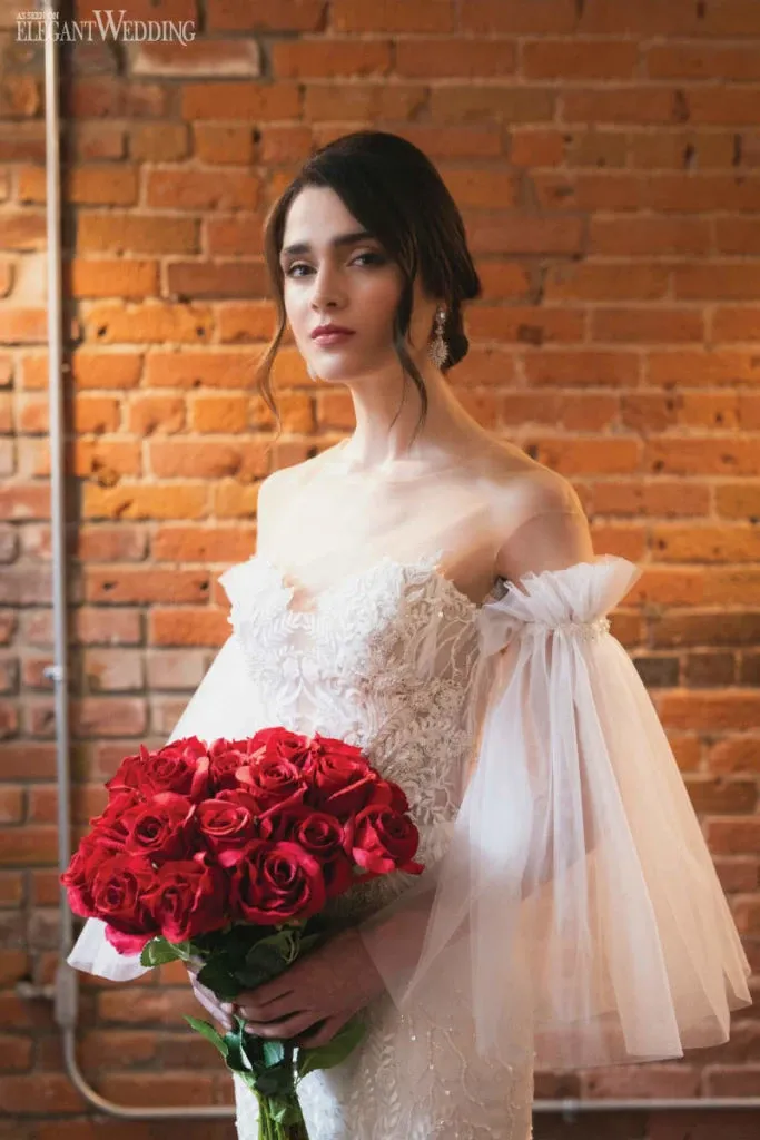 A bride stands elegantly against a rustic brick wall, showcasing her off-the-shoulder, lace bridal gown with sheer, flowing sleeves. She holds a vibrant bouquet of red roses, adding a pop of color to the scene. Her hair is styled in a classic updo, adorned with subtle, elegant earrings. This image represents the wedding beauty services of Studio Caroline Théoret, featured on Mariage Elegant, Canada.