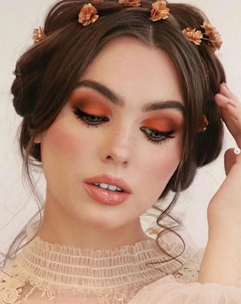 In this image, a bride showcases an elegant beauty look crafted by Studio Caroline Théoret, featured under the Wedding Beauty category on Mariage Elegant, Canada. The bride is adorned with a sophisticated updo accented by small floral hairpieces, perfectly complementing her soft, romantic makeup. Her eye makeup features warm, earthy tones with a bold copper eyeshadow, coupled with voluminous lashes that enhance her eyes. Her cheeks are delicately blushed, and her lips have a glossy peach tint. She wears a lace bridal gown with intricate detailing visible at the neckline, embodying a classic bridal elegance.