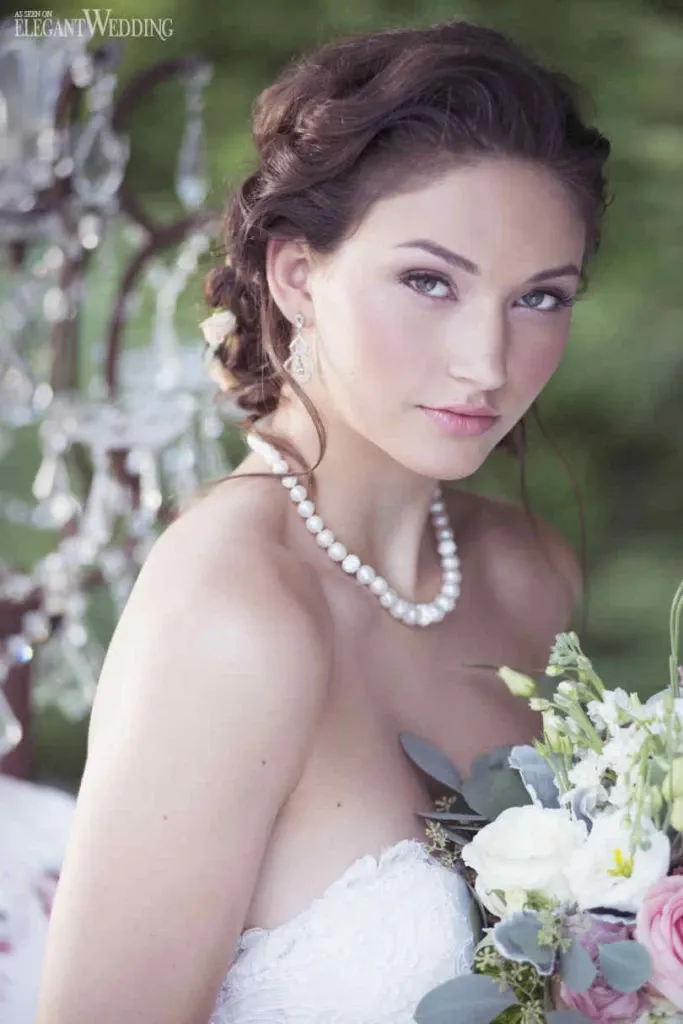 The image features a bride with an elegant updo adorned with delicate floral accents, showcasing the work of Studio Caroline Théoret in the Wedding Beauty category. She is wearing a classic pearl necklace and drop earrings, complementing her natural makeup look. The bride is holding a bouquet of pastel-colored flowers, including white roses and greenery. The soft-focus background features a decorative crystal chandelier, adding to the romantic ambiance. This image highlights the artistry of bridal beauty suitable for weddings.