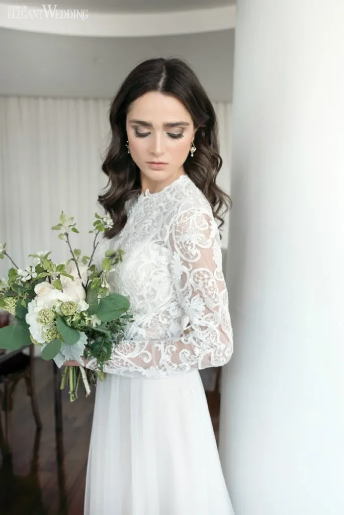 A bride stands gracefully, showcasing her intricate lace wedding gown with long sleeves and a flowing skirt. She holds a lush bouquet of white and green flowers, complementing her elegant look. Her dark hair cascades in soft waves, and she gazes downward, highlighting her delicate earrings. This beautiful bridal style is expertly crafted by Studio Caroline Théoret, featured in the Wedding Beauty category on Mariage Elegant, Canada. The backdrop of sheer white curtains adds to the serene and sophisticated ambiance, making this image a perfect representation of refined wedding beauty.