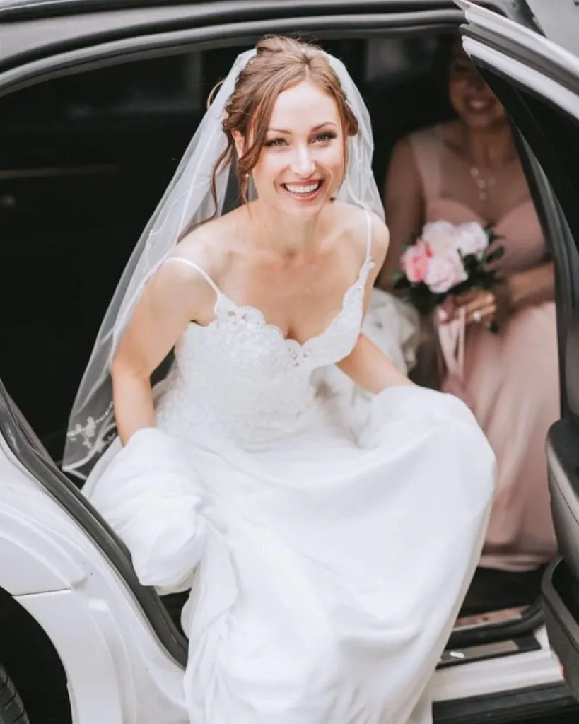 A bride wearing a lace wedding dress and veil smiles brightly as she steps out of a car, assisted by a person in the background holding a bouquet of pink and white flowers. This image showcases the exquisite bridal beauty services provided by Studio Caroline Théoret, under the Wedding Beauty category on Mariage Elegant, Canada. The bride’s elegant look is a testament to the professional wedding beauty expertise offered by this vendor.