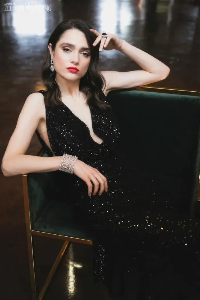 A bride sits elegantly on a dark green velvet chair in a dimly lit room, exuding sophistication. She is wearing a stunning black sequined gown that drapes beautifully to the floor. The bride's hair is styled in soft waves, complementing her bold red lipstick and expertly applied makeup, which highlights her features with precision. Her look is accessorized with sparkling statement earrings, a chunky bracelet, and a striking black ring, adding a touch of glamour. This image showcases the wedding beauty expertise of Studio Caroline Théoret, highlighting their ability to create a chic and elegant bridal look.