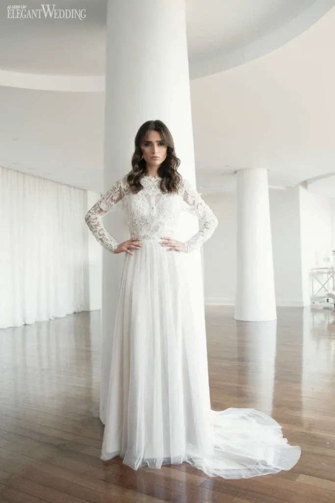 A bride stands in a beautifully lit room with large white columns and a glossy wooden floor, wearing an elegant, long-sleeved lace wedding dress designed by Studio Caroline Théoret. Her hair is styled in soft waves, and she exudes a serene and confident presence. This image, featured in the Wedding Beauty category on Mariage Elegant, Canada, captures the exquisite craftsmanship and attention to detail that Studio Caroline Théoret is known for in wedding attire and beauty styling.