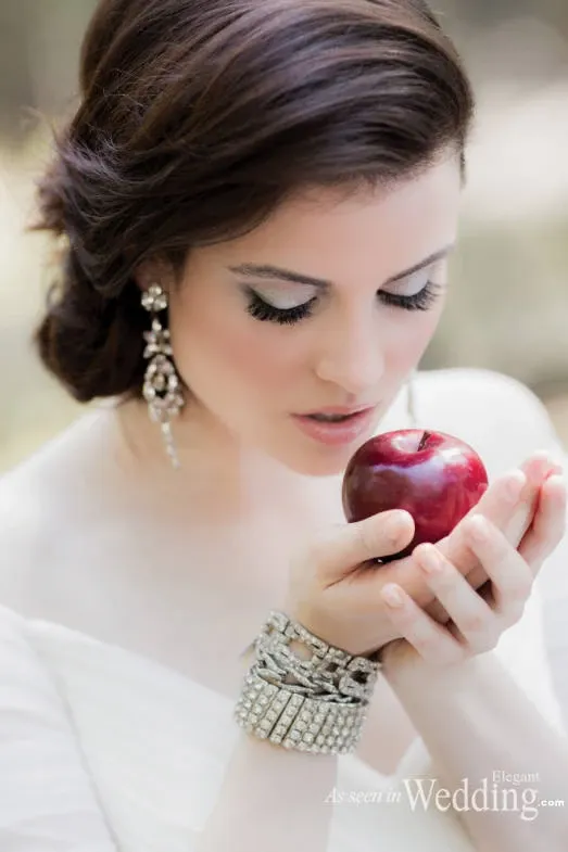In this elegant image, a bride holds a deep red apple delicately in both hands, reflecting a fairy-tale theme. She has an intricate bridal hairstyle and dramatic makeup, accentuating her expressive eyes. Her attire features a pure white, off-the-shoulder gown accompanied by sparkling, statement earrings and a bracelet adorned with crystals. The soft, blurred background provides a serene, natural setting. This captivating bridal look is crafted by Studio Caroline Théoret, highlighting their expertise in the Wedding Beauty category for Mariage Elegant, Canada.