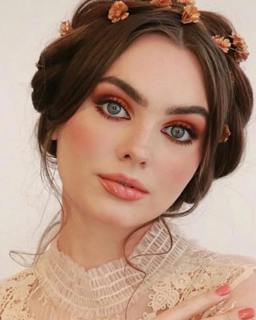 The image features a bride showcasing elegant, professionally styled hair and makeup by Studio Caroline Théoret in the Wedding Beauty category. Her dark hair is styled in an intricate updo adorned with delicate golden floral accessories that complement her warm, sophisticated makeup. Her eyes are highlighted with rich, earthy tones, and her lips are a soft, glossy nude. She is wearing an intricately designed lace dress, adding to the romantic and refined wedding aesthetic. This beautifully crafted look exemplifies the services offered by Studio Caroline Théoret for brides seeking exceptional beauty styling in Canada.