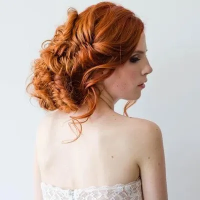 A bride with vibrant, cascading red hair styled in an intricate updo, captured in a side profile against a simple, light background. Her elegant hairstyle, showcasing expertly crafted braids and curls, exemplifies the expertise of Studio Caroline Théoret in the Wedding Beauty category. She wears a strapless lace dress, highlighting a classic and sophisticated bridal look, perfect for a modern wedding.