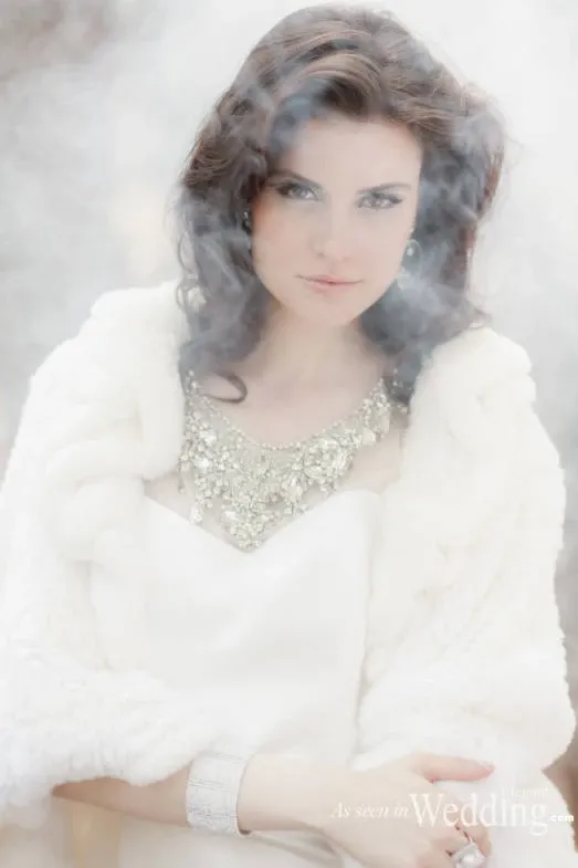 A bride gazes softly into the camera, enveloped in a dreamy ambiance created by surrounding mist. She wears a luxurious, white faux fur stole, highlighting her elegant wedding gown adorned with intricate beadwork around the neckline. Her dark, wavy hair frames her face, complemented by subtle yet sophisticated makeup. This image represents the exquisite work of Studio Caroline Théoret in the Wedding Beauty category, showcasing their ability to create ethereal and timeless bridal looks.