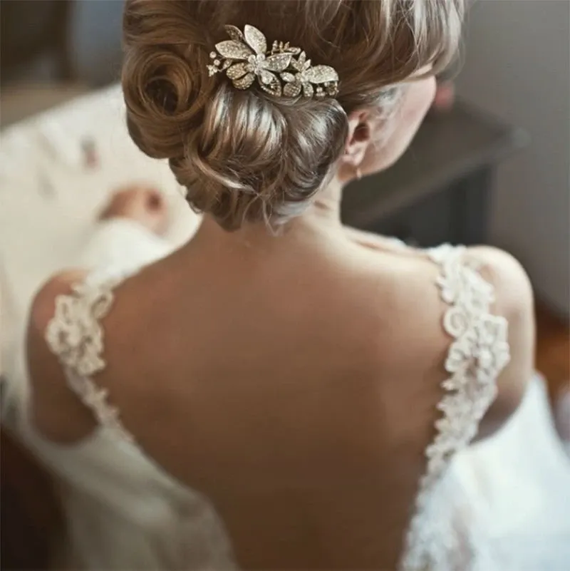 This image showcases a bride from behind, styled by Studio Caroline Théoret in the Wedding Beauty category. Her hair is elegantly arranged in a low bun, adorned with a sparkling, intricately designed hairpiece featuring leaf and floral motifs. The bride's dress features delicate lace straps and an open back, adding a touch of sophistication and romance. The setting appears to be a softly lit dressing area, emphasizing the serene and intimate preparation moments before the wedding ceremony.