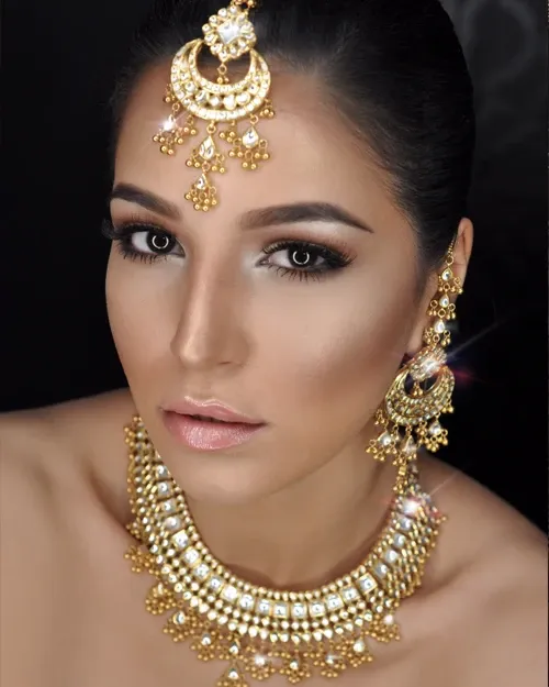 A bride showcases exquisite bridal makeup and jewelry, highlighting the services of Amber Esthetics in the Wedding Beauty category. The image features a woman with elegantly contoured makeup, including striking eye makeup and soft, luminous skin tones. Her look is complemented by ornate gold jewelry, including a statement necklace, chandelier earrings, and a matching maang tikka, each adorned with intricate detailing and sparkling gemstones. This image reflects the high-quality beauty services available for brides at Mariage Elegant in Canada.