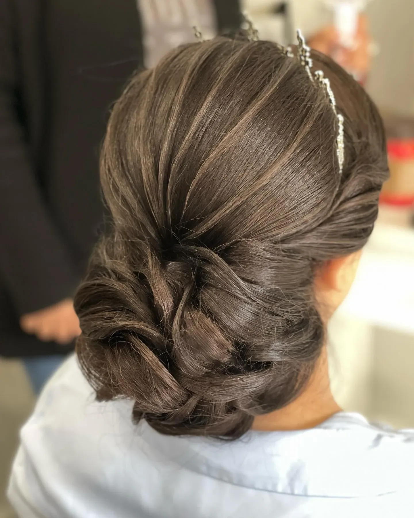 A bride with elegantly styled dark brown hair sits in a beauty salon, showcasing a sophisticated updo adorned with delicate hair accessories. The hairstyle is meticulously crafted, featuring intricate twists and a smooth finish, perfect for a wedding day. This image represents the exceptional services offered by Amber Esthetics in the Wedding Beauty category, highlighting their expertise in bridal hairstyling. The setting suggests a professional environment dedicated to wedding preparations, contributing to an unforgettable celebration.