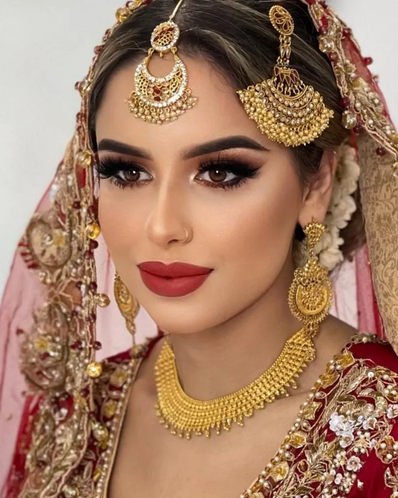 A bride with elaborate wedding makeup and jewelry, showcasing the work of Amber Esthetics in the Wedding Beauty category. The bride wears a striking red lipstick, dark, dramatic eye makeup, and bold, perfectly shaped eyebrows. Her hair is styled elegantly, with a delicate headpiece featuring intricate gold details and adorned with small pearls, matching the decorative maang tikka on her forehead. She is dressed in an ornate red and gold bridal outfit, reflecting traditional wedding attire. The look is completed with an exquisite gold necklace and large, detailed earrings, exemplifying luxurious bridal beauty offerings by Amber Esthetics in Canada.