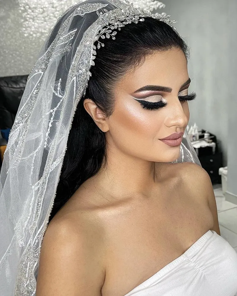 A bride showcasing a stunning makeup look by Amber Esthetics, a vendor in the Wedding Beauty category at Mariage Elegant, Canada. She has elegantly styled dark hair adorned with a sparkling, jeweled headpiece and a delicate lace veil. Her makeup features dramatic winged eyeliner, long lashes, and expertly contoured cheeks, complemented by a soft pink lip color. The bride's serene expression highlights the professional artistry of Amber Esthetics, emphasizing the vendor's expertise in enhancing bridal beauty. The backdrop is a softly lit, elegant setting, suitable for wedding preparations.