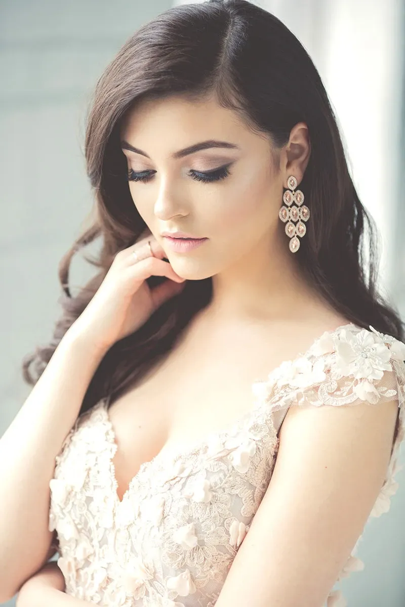 A bride with long, dark hair styled in soft waves looks down gracefully. She is wearing elegant, floral lace earrings and a delicate, embroidered wedding gown with floral appliqués on the straps. The makeup is flawlessly applied, highlighting her eyes and lips, showcasing the expertise of Amber Esthetics. This image is part of the Wedding Beauty category for Mariage Elegant, Canada, emphasizing the refined and sophisticated bridal look offered by the vendor.