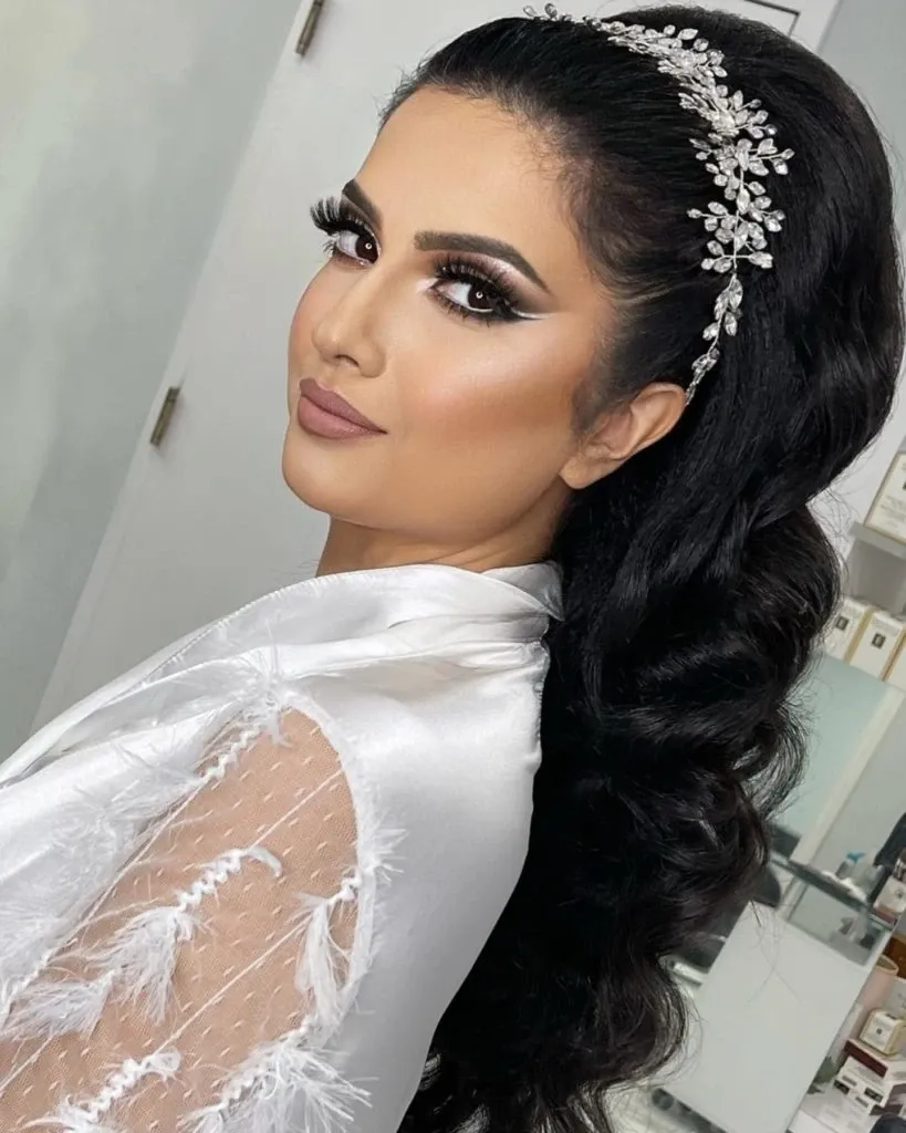 In this image, a bride is elegantly styled by Amber Esthetics, showcasing their expertise in the Wedding Beauty category. The bride's hair is styled in voluminous curls cascading down her back, adorned with an intricate, sparkling headpiece that adds a touch of glamour. Her makeup is expertly applied, featuring dramatic winged eyeliner and soft, neutral lip color, enhancing her natural beauty. She wears a delicate, white lace robe, perfect for a wedding preparation setting. The soft, serene background complements the focus on her sophisticated bridal look by Amber Esthetics.