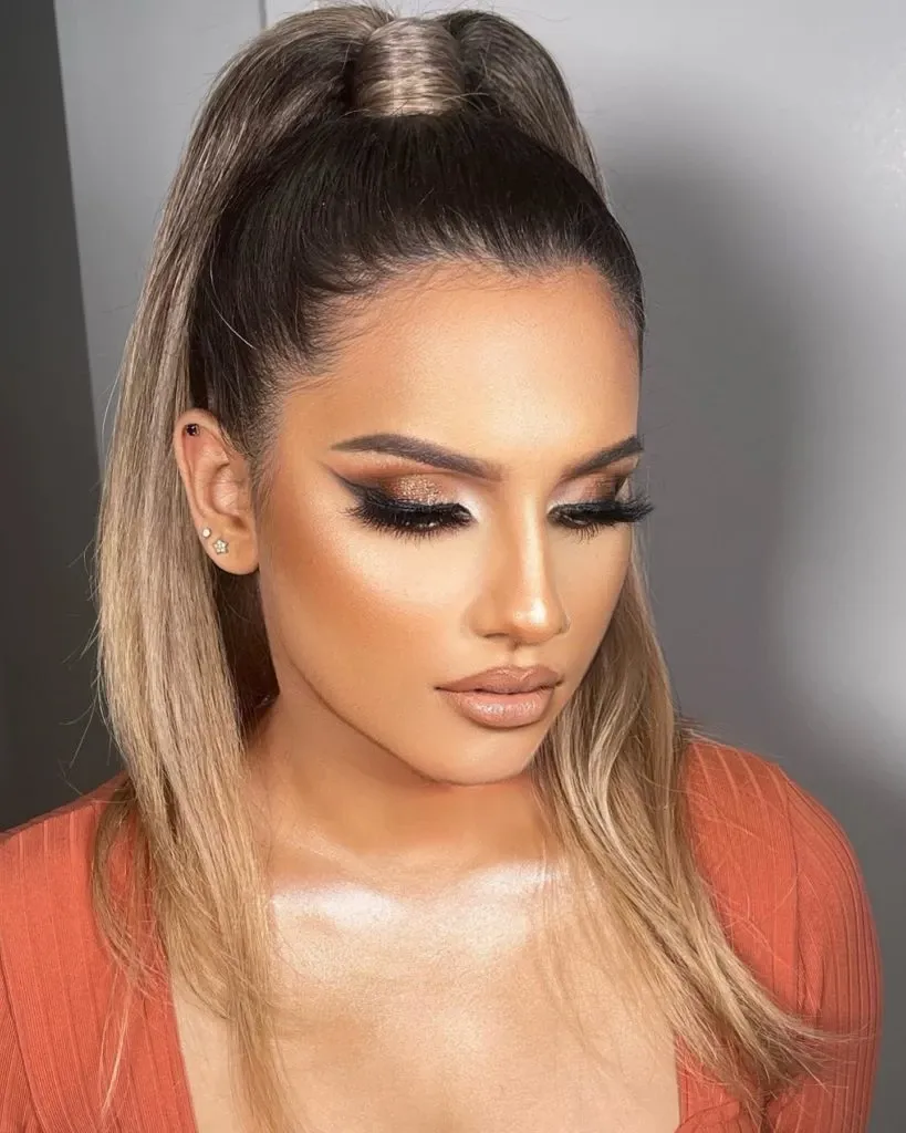 This image features a woman with expertly applied makeup by Amber Esthetics, highlighting their skills in the Wedding Beauty category. The woman has long, smooth brown-blonde hair styled in a high ponytail, emphasizing her elegant features. Her makeup includes bold, arched eyebrows, dramatic winged eyeliner, lush eyelashes, and warm-toned eyeshadow, complemented by a subtle contour and a nude lip color. She is wearing a rust-colored top that adds a touch of sophistication, making this look ideal for a wedding ceremony or reception. Amber Esthetics showcases their expertise in providing stunning bridal makeup services, perfect for a memorable wedding day.