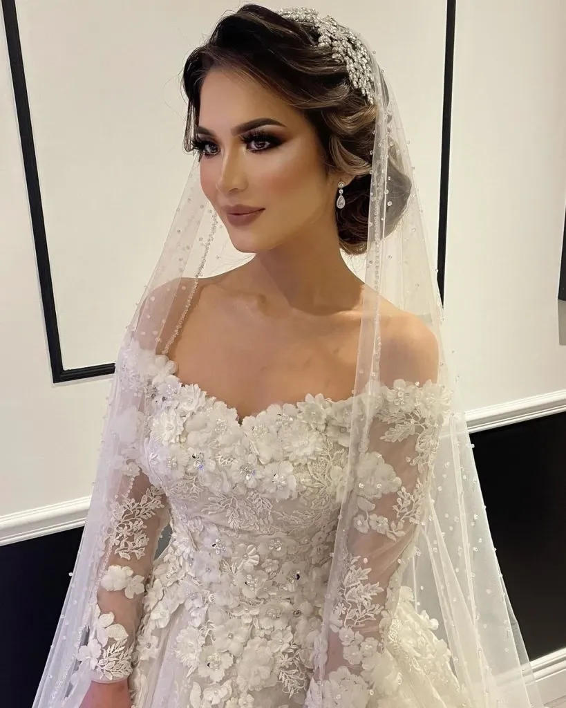A bride beautifully prepared by Amber Esthetics, featured in the Wedding Beauty category on Mariage Elegant, Canada. The bride is wearing a stunning off-the-shoulder lace gown adorned with floral appliqués and subtle beading, perfectly complemented by a delicate veil embellished with tiny pearls. Her makeup is expertly done, highlighting her features with soft, natural tones. The hairstyle is an elegant updo, decorated with a sparkling hair accessory, enhancing the bridal look. The background is minimal, with elegant black and white decor that ensures all focus remains on the bride.