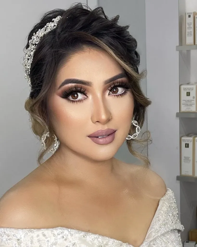 A bride with elegant makeup and a stylish updo, featuring subtle highlights, is shown in this image. She is wearing a delicate, beaded hair accessory and sparkling drop earrings. Her makeup consists of expertly blended eyeshadow, long lashes, and a soft pink lip color, highlighting her refined beauty. The background includes shelves with beauty products, suggesting a professional studio setting. This image represents Amber Esthetics, a vendor in the Wedding Beauty category, as featured on Mariage Elegant in Canada.
