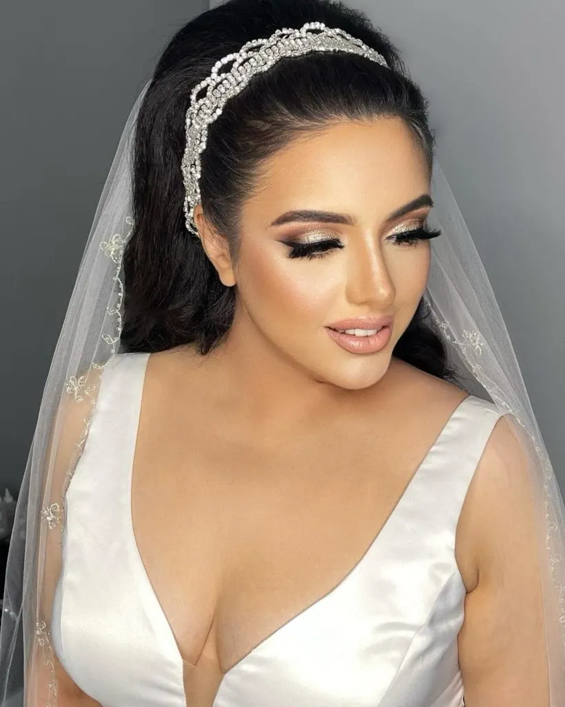 In this image from Mariage Elegant's wedding vendor listing, Amber Esthetics showcases their expertise in the Wedding Beauty category. The bride is elegantly styled with expertly applied makeup featuring soft, smoky eyes and perfectly contoured cheeks. Her lips are painted in a natural nude shade, complementing her flawless complexion. She wears a stunning white gown with a deep V-neckline and is adorned with a delicate, beaded headband that secures her tulle veil. The hairstyle is a sophisticated updo, adding to the bridal elegance captured in the photo.