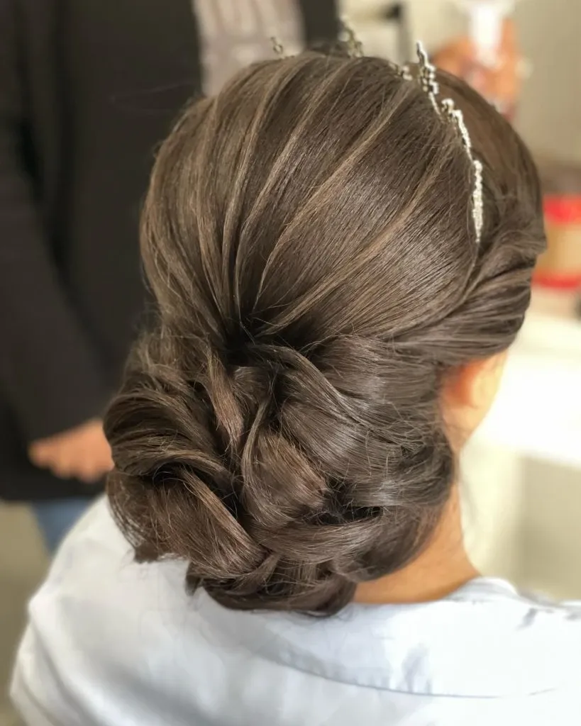 A bride showcases an elegant and intricately styled updo crafted by Amber Esthetics under the wedding beauty category. Her dark brown hair is styled into a smooth, twisted bun with delicate strands forming a voluminous, sophisticated look. A sparkling hairpiece is subtly placed, adding a touch of glamour to the overall style, perfect for a wedding day celebration. The background features a blurry setting with a hint of a person holding a drink, emphasizing the focus on the exquisite hairstyle by this esteemed wedding beauty vendor in Canada.