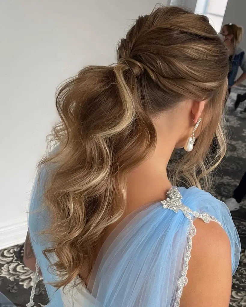 A woman with elegantly styled wavy hair is showcased in the image, highlighting the expert work of Amber Esthetics in the Wedding Beauty category. Her hair is skillfully pinned back into a sophisticated half-up hairstyle, allowing the loose curls to cascade down gracefully. She is wearing a delicate blue gown adorned with a jeweled brooch on the shoulder, perfect for a wedding setting. The soft lighting and intricate details emphasize the luxurious and refined style suitable for bridal occasions.