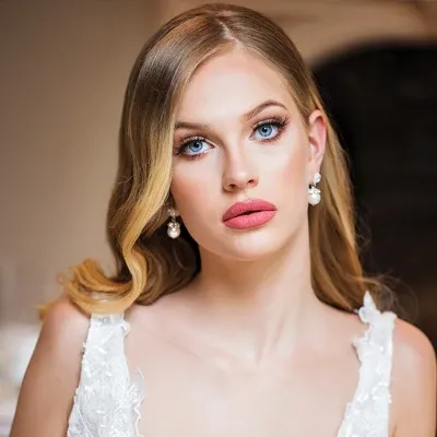 A bride with flawless makeup and elegant hairstyle is showcasing her look, embodying the services provided by Amber Esthetics in the Wedding Beauty category. Her makeup features bold, rosy lipstick and expertly applied eye makeup that highlights her blue eyes. She wears her blonde hair in soft waves, complemented by pearl earrings. The detailed lace straps of her wedding gown add a touch of sophistication, reflecting the high-quality bridal beauty services offered by Amber Esthetics at Mariage Elegant in Canada.