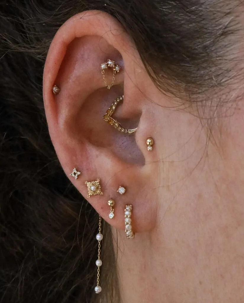 Le gros plan montre une oreille ornée de plusieurs bijoux délicats, comprenant des anneaux et des clous en or, certains décorés de petites perles. Ces pièces élégantes et sophistiquées, proposées par Paradise Montreal, s'intègrent parfaitement dans la catégorie Beauté pour le mariage, ajoutant une touche de glamour et de raffinement, idéale pour une mariée désirant se démarquer lors de son mariage.
