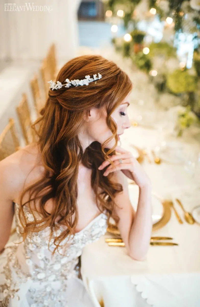 The image showcases a bride with long, wavy, auburn hair adorned with a delicate floral hairpiece, styled by Salon Deauville under the Wedding Beauty category. She is seated at an elegantly set table, thoughtfully gazing downward. Her intricate lace wedding gown adds to the sophisticated ambiance. The background features blurred greenery and warm lighting, enhancing the romantic and luxurious atmosphere typical for wedding events.