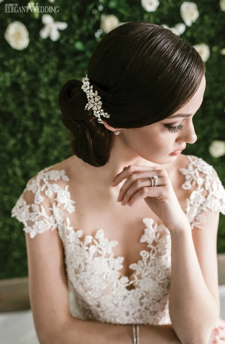A bride with elegantly styled brown hair adorned with a delicate, floral hairpiece gazes downward in a thoughtful pose. She wears a stunning white lace wedding gown, featuring intricate floral detailing on the shoulders and sleeves. The background showcases a lush green wall with white roses, adding a touch of natural beauty. The image represents the services of Salon Deauville in the Wedding Beauty category, highlighting their expertise in bridal styling and elegance for Mariage Elegant in Canada.