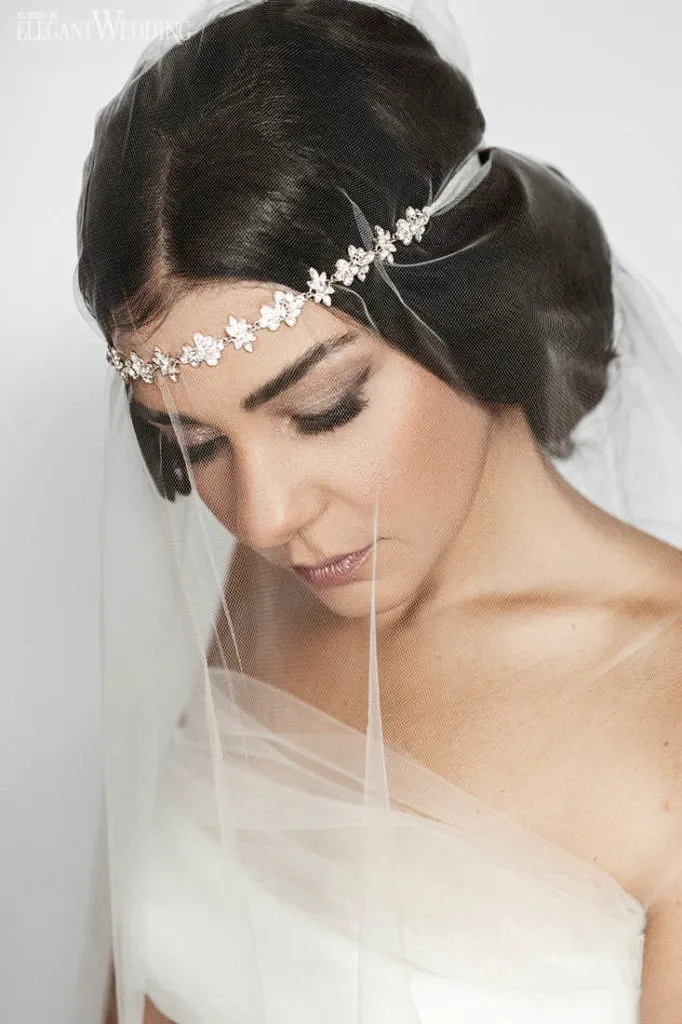 In the image, a bride is beautifully styled by Salon Deauville, featured in the Wedding Beauty category on Mariage Elegant, Canada. The bride has a sophisticated, low bun and is wearing a delicate, floral headband adorned with crystals that elegantly complements her veil. Her makeup is classic and refined, emphasizing natural tones and enhancing her features for a radiant bridal look. The image captures the essence of elegance and attention to detail, showcasing the salon's expertise in wedding beauty preparations.