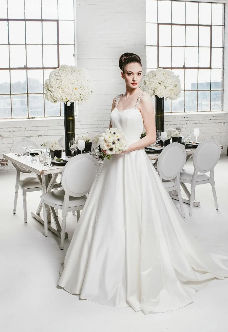 A bride stands elegantly in a chic, modern setting, showcasing her stunning white gown. Her hair and makeup, expertly crafted by Salon Deauville, exemplify the highest standards of wedding beauty. She holds a bouquet of white flowers, complementing her sophisticated look. The room is beautifully arranged with a reception table set with white and black tableware, surrounded by white chairs. Large vases filled with lush white floral arrangements add to the ambiance, capturing the essence of a refined and elegant wedding celebration in the heart of Canada.