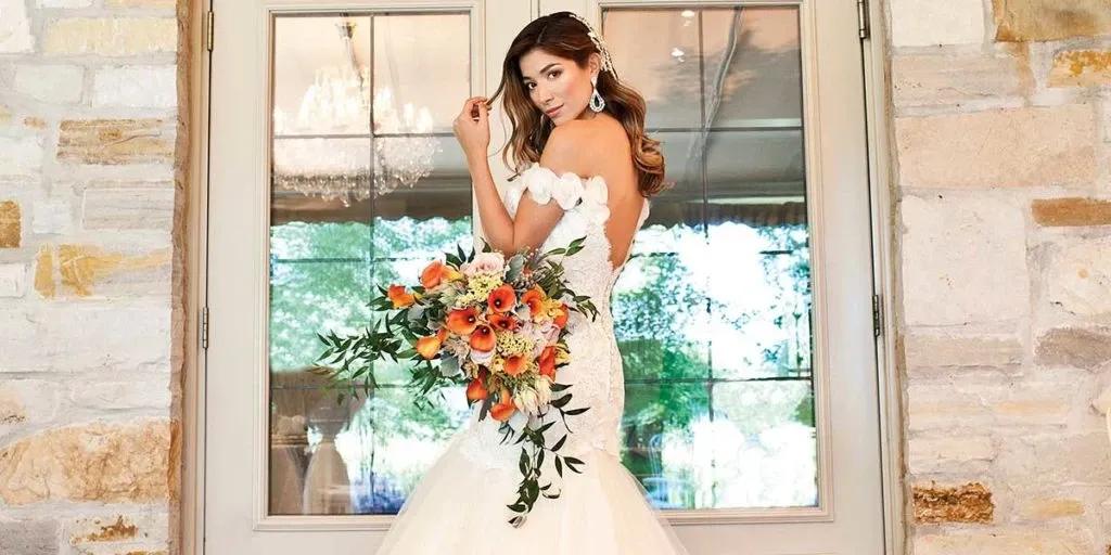 A bride stands elegantly in front of a large window, wearing a stunning, off-the-shoulder wedding gown that features intricate lace details. She holds a vibrant bouquet of orange and white flowers with lush greenery. Her hair is styled in loose waves, and she wears delicate earrings that complement her sophisticated look. The setting exudes a luxurious atmosphere, enhanced by the visible chandelier inside. This image highlights the beauty services offered by Salon Deauville, a prominent vendor in the Wedding Beauty category on Mariage Elegant, Canada.