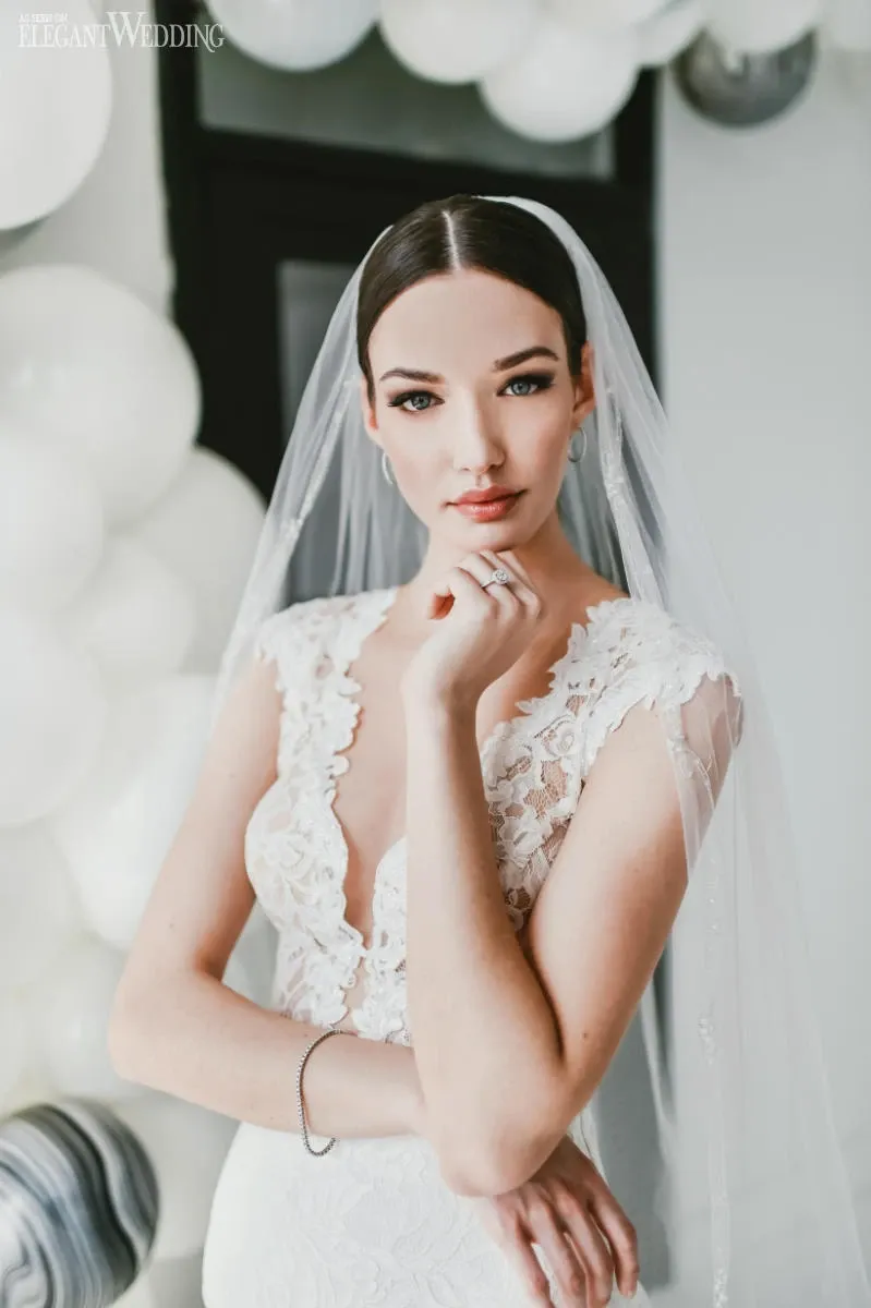 In this image from Mariage Elegant, Canada, a bride is elegantly prepared by Salon Deauville, a leading vendor in the Wedding Beauty category. The bride is wearing a stunning white lace wedding gown with intricate detailing and a classic veil, perfectly complementing her sophisticated makeup and sleek, dark hair styled in an updo. The serene expression on her face and the gentle pose of her hand near her chin add an air of grace and poise. In the background, white balloons create a soft and celebratory atmosphere, highlighting the beauty and elegance of this special wedding moment.