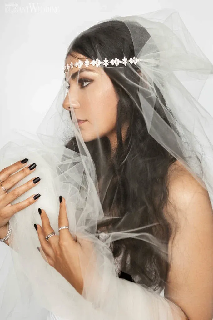The image features a bride with long, dark, wavy hair styled elegantly, showcasing the expertise of Salon Deauville in the wedding beauty category. She is wearing a delicate, floral headband that complements the sheer veil draped over her shoulders and arms. Her makeup is subtle, enhancing her natural beauty, with an emphasis on striking eyeliner and soft lipstick. Her nails are painted black, adding a modern touch to her look. This image captures the refined elegance and attention to detail that Salon Deauville offers to brides in Canada, as featured on the Mariage Elegant wedding planning and vendor listing site.
