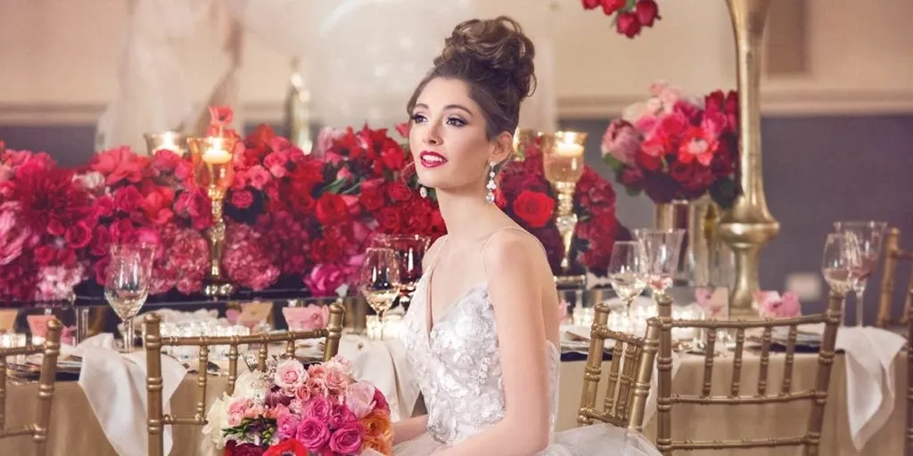 A bride with an elegant updo and dramatic makeup, showcasing beauty services by Salon Deauville, is seated at a lavishly decorated wedding reception table. The setting features an array of vibrant red and pink floral arrangements, reflecting a romantic theme. Gold chiavari chairs and candle-lit glassware enrich the luxurious atmosphere, highlighting the salon's expertise in wedding beauty.