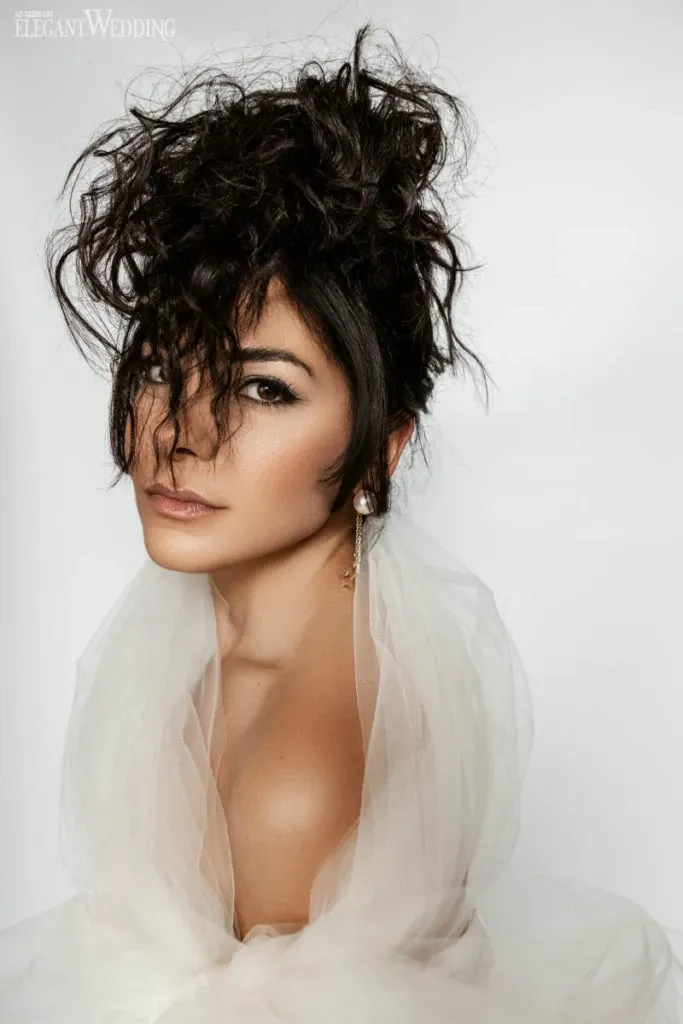 In this elegant portrait, a woman with an intricate, curly updo styled by Salon Deauville is featured, highlighting the vendor’s expertise in wedding beauty. The hair is artfully arranged, creating a voluminous and sophisticated look perfect for a wedding day. Her makeup is expertly applied, emphasizing her eyes and giving her a refined, glamorous appearance. She is wearing luxurious white fabric draped around her shoulders, contributing to the overall bridal theme. This image exemplifies Salon Deauville's skill in creating exquisite bridal styles suitable for Mariage Elegant's wedding planning and vendor listing in Canada.