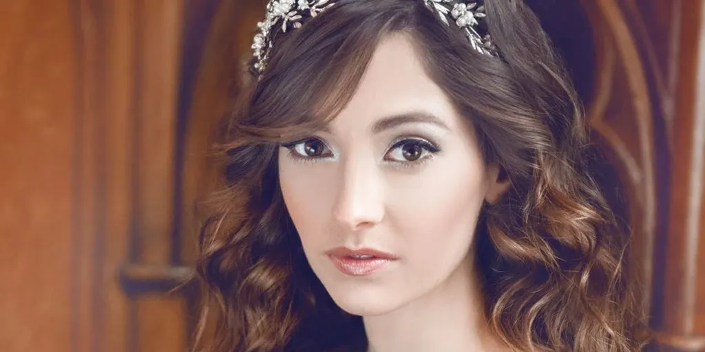 In this image, a woman is elegantly dressed, showcasing a stunning bridal hairstyle by Salon Deauville, featured under the Wedding Beauty category. Her long, wavy hair cascades beautifully, adorned with an intricate floral headband adding a touch of sophistication. The soft, natural makeup complements her look, emphasizing her eyes and enhancing her features. The background suggests a luxurious setting, ideal for a wedding environment, highlighting the expertise of Salon Deauville in creating exquisite bridal styles.