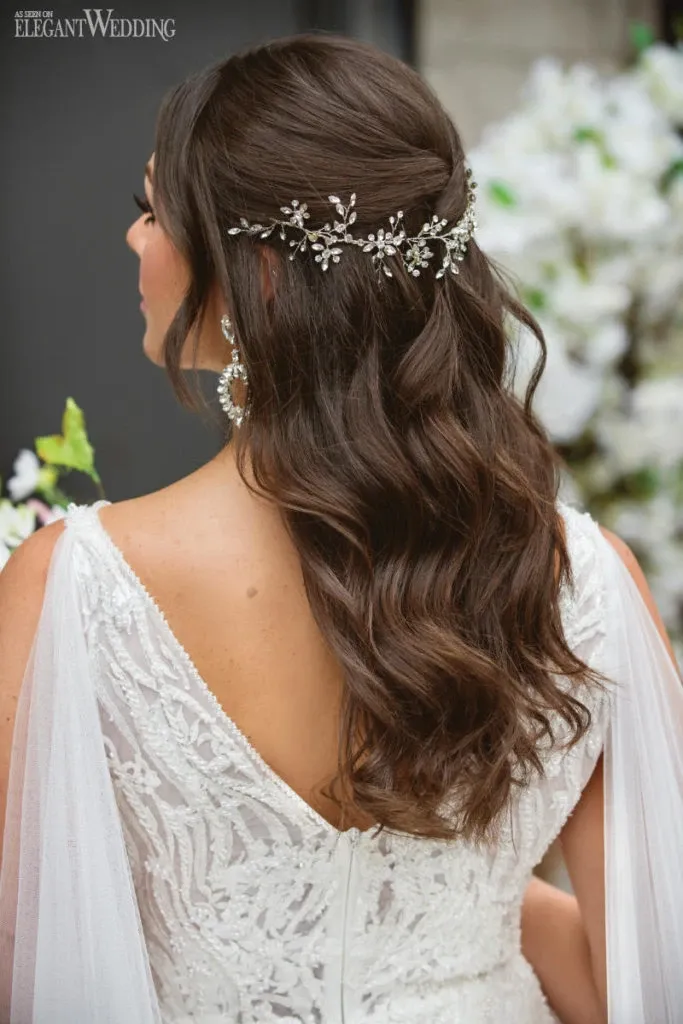 A bride with elegantly styled brown hair, featuring soft waves cascading down her back, is adorned with a delicate jeweled hairpiece at the top. She is wearing a white lace wedding gown with intricate detailing on the back and sheer fabric at the shoulders. The background includes lush, green foliage and white flowers, adding a natural touch to the setting. This image showcases the bridal beauty services offered by Salon Deauville, categorized under Wedding Beauty, highlighting their expertise in creating beautiful wedding hairstyles for brides.