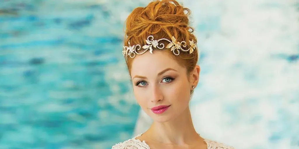 A bride with elegant auburn hair styled in an intricate updo adorned with a delicate, jeweled headpiece gazes softly at the camera. She is wearing a white lace gown with intricate detailing, highlighting her sophisticated look. The background features a blurred, blue-toned artistic design, complementing the elegance of the scene. This image showcases the expertise of Salon Deauville in the Wedding Beauty category, emphasizing their talent in creating stunning bridal hairstyles and appearances for weddings.