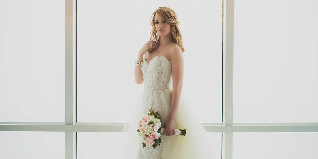 A bride stands elegantly in a bright, minimalist setting, showcasing a strapless lace wedding gown. She holds a bouquet of soft pink and white flowers, adding a touch of color to the scene. Her hair is styled in loose, flowing waves, and she wears subtle, polished makeup, highlighting her natural beauty. This image, featuring Salon Deauville, represents the Wedding Beauty category on Mariage Elegant, Canada, capturing the essence of bridal elegance and style.