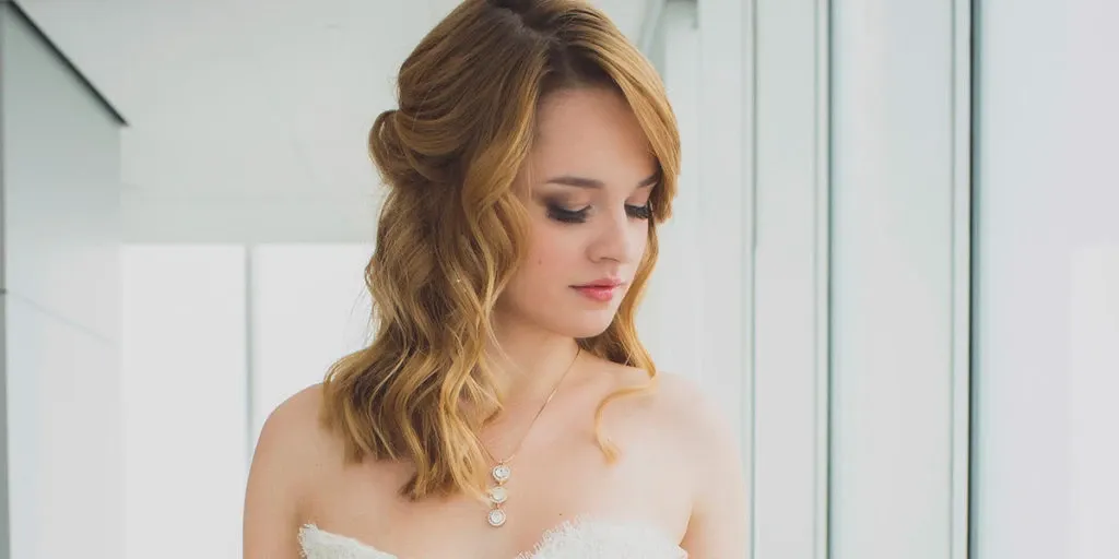 A bride with wavy, shoulder-length blonde hair and professionally styled makeup by Salon Deauville poses near a large, bright window. She wears an elegant strapless white gown with delicate lace details and a layered pendant necklace. This image highlights the Wedding Beauty expertise of Salon Deauville, showcasing their skill in creating a classic bridal look for weddings in Canada.