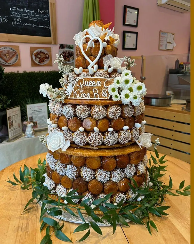 The image features an exquisite wedding cake by vendor AliceÔchoux, categorized under Wedding Cakes on Mariage Elegant, Canada. The cake is a multi-tiered croquembouche, elegantly adorned with cream-filled profiteroles covered in caramelized sugar. Delicate white flowers, including roses and daisies, are artistically arranged around the layers, adding a touch of elegance. A decorative heart topper with the words 'Queen Kma B & A' is placed prominently at the top, suggesting personalization for a special occasion. The base of the cake is surrounded by vibrant green foliage, providing a fresh and natural look. The backdrop includes a glimpse of certificates and awards on the wall, indicative of the vendor's professional recognition and expertise in wedding cake design.