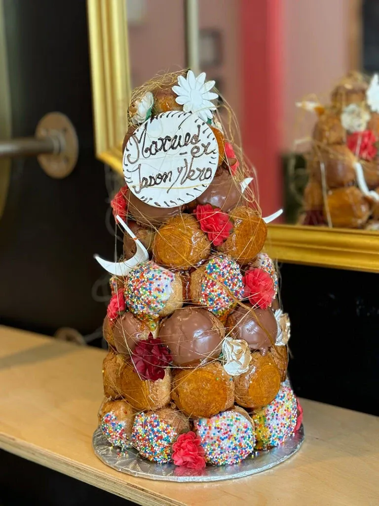 This image showcases a stunning croquembouche created by AliceÔchoux, featured in the Wedding Cakes category on Mariage Elegant, Canada. The croquembouche is elegantly arranged with a variety of cream-filled profiteroles, some are coated in chocolate and others are covered in colorful sprinkles. Red and white decorative flowers add a touch of sophistication, and a delicate spun sugar netting encases the structure. At the top, a personalized plaque with the inscription 'Marcus & Jason' in elegant script provides a personal touch, perfect for a wedding celebration. The reflection in the mirror behind the dessert highlights the intricate details and craftsmanship.