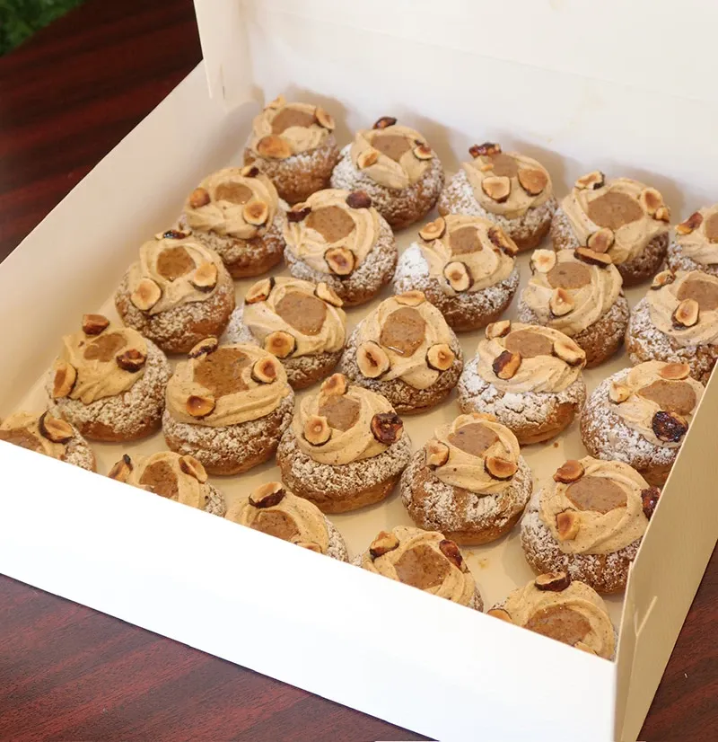 This image features a box of meticulously crafted wedding pastries by AliceÔchoux, categorized under Wedding Cakes at Mariage Elegant, Canada. The pastries are neatly arranged in a white box, each topped with a generous layer of creamy frosting and garnished with chopped nuts. The light dusting of powdered sugar adds an elegant touch, making these treats ideal for sophisticated wedding celebrations. The wooden table beneath the box complements the luxurious presentation of these wedding confections.