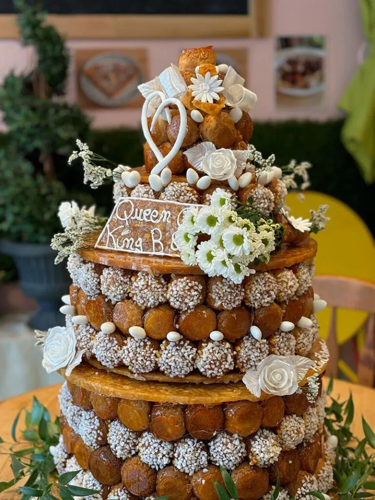 This image showcases a stunning and elaborate wedding cake crafted by AliceÔchoux, under the wedding cakes category for Mariage Elegant in Canada. The cake is a grand three-tiered croquembouche adorned with delicate white flowers and intricate sugar decorations, including roses and dainty heart toppers. Each profiterole is evenly coated with sugar crystals, creating a textured, elegant look. Greenery surrounds the base, adding a natural touch to the sophisticated design. A small inscription on one tier reads 'Queen of Kora B.' indicating a possible personalized message. This masterpiece exemplifies the art of wedding cake design.