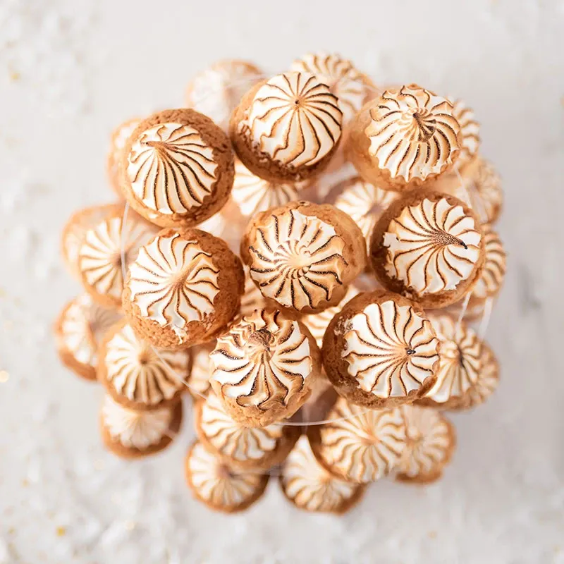 This image showcases an elegant display of beautifully crafted wedding pastries by AliceÔchoux, part of the Wedding Cakes category on Mariage Elegant, Canada. The photo features a cluster of choux pastries, artistically arranged in a tower. Each pastry is topped with delicately piped, golden-brown meringue, highlighting intricate patterns against a soft, white background, creating a visually appealing centerpiece perfect for a sophisticated wedding celebration.