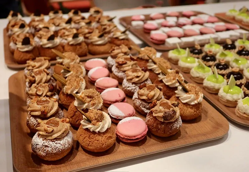 The image showcases an exquisite display of wedding desserts created by AliceÔchoux, a renowned vendor in the Wedding Cakes category at Mariage Elegant, Canada. The arrangement features a variety of intricately designed pastries and macarons. In the foreground, pink macarons are artfully paired with cream-filled pastries topped with chocolate accents and nuts, presented on a wooden platter. In the background, other platters display an assortment of confections, including bright green glazed pastries and bite-sized treats, creating a visually appealing assortment perfect for a sophisticated wedding reception.
