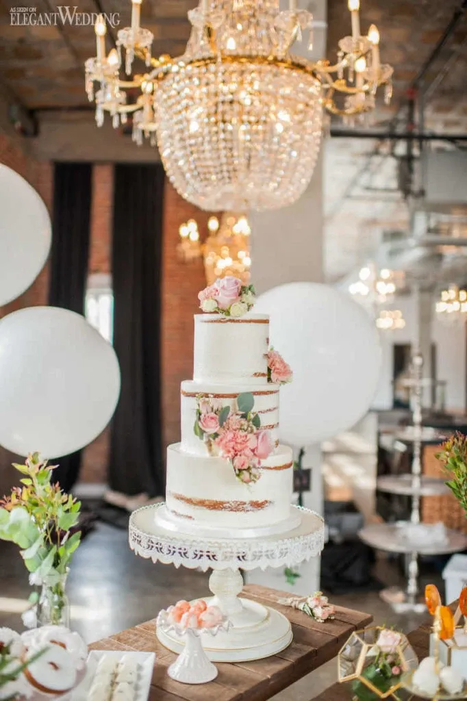 This image showcases a stunning wedding cake crafted by Pâtisserie Montebello, under the category of Wedding Cakes from Mariage Elegant, Canada. The cake is a three-tiered masterpiece, elegantly adorned with delicate pink roses and greenery, which gracefully cascade down the sides. It rests on an intricately detailed white cake stand. The background reveals the luxurious setting of a wedding venue, characterized by a grand crystal chandelier and exposed brick walls, creating an ambiance of rustic elegance. Additional decorative elements, such as delicate floral arrangements and soft lighting, enhance the sophisticated atmosphere, making this an ideal choice for a stylish wedding celebration.